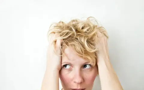 Kobieta z blond włosami wplata dłonie we włosy, jej wyraz twarzy sugeruje wahania nastroju i niepokój.