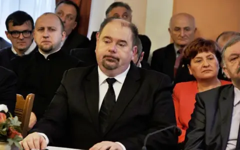 Jarosław Paluch w czarnym garniturze siedzi przy stole, otoczony innymi osobami.