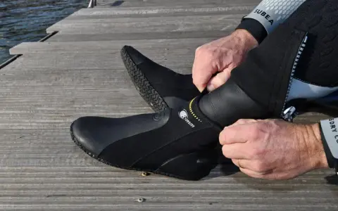 Mężczyzna zakłada czarne buty neoprenowe na drewnianym pomoście przy wodzie.