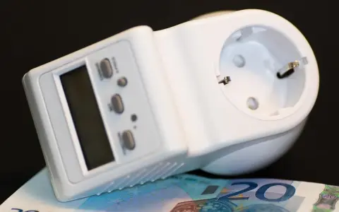 Biały miernik zużycia prądu z wyświetlaczem i przyciskami, leżący na banknocie 20 euro.