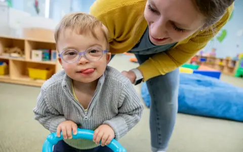 Chłopiec z zespołem Downa, w okularach, z językiem na brodzie, bawi się z terapeutką. To przykład pracy w obszarze oligofrenopedagogiki.