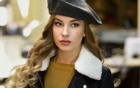Elegancka kobieta w grafitowym beretcie, pokazująca, jak się nosi beret, z burzą blond loków i w stylowej kurtce.