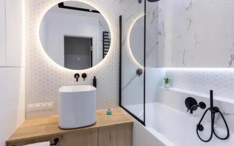 Elegante bagno senza finestra con lavabo bianco, specchio rotondo illuminato e doccia con finiture nere.