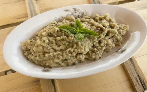 Hummus di melanzane cremoso, guarnito con basilico fresco, servito in un piatto ovale su un tavolo di legno grezzo.