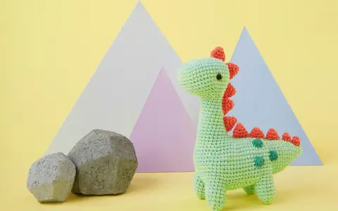 Adorable dinosaurio crochet verde con púas naranjas y detalles verdes. Dos rocas grises y formas geométricas de colores pastel al fondo.