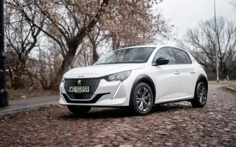 Biały Peugeot e-208 na brukowanej ulicy, z drzewami w tle.