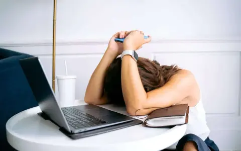Personne épuisée, tête sur les bras, téléphone à la main, devant un ordinateur portable. L'effet écran de la fatigue est palpable.