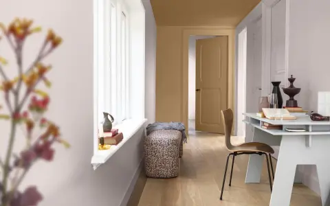 Ett rum med ljusa väggar och en dörr som är målad i en varm, gyllene ton. En vit fönsterbräda med böcker och en vas. En bänk med blommigt tyg och en trästol vid ett skrivbord.