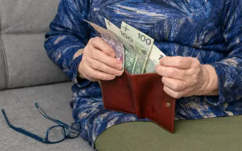 Dłoń starszej osoby wkłada pieniądze do portfela. Widać banknoty 100 zł. To wsparcie dla osób niepełnosprawnych, jak 500 plus dla niepełnosprawnych.