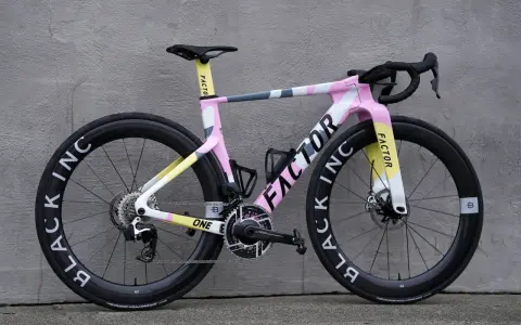 A pink, yellow, and gray Factor road bike with black wheels against a gray wall.