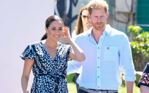 Meghan och Harry ler glatt under ett besök utomhus. Meghan bär en mönstrad klänning och Harry en ljusblå skjorta.