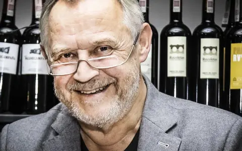 Marek Kondrat, z uśmiechem i okularami, na tle butelek wina. Jego majątek to nie tylko sukcesy zawodowe, ale i pasja do trunków.
