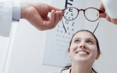 Okulista trzyma okulary przed twarzą uśmiechniętej kobiety na tle tablicy Snellena.