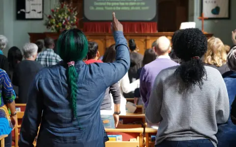 A woman with green hair raises her hand, perhaps inspired by the lyrics on screen, offering chant ideas for the congregation.