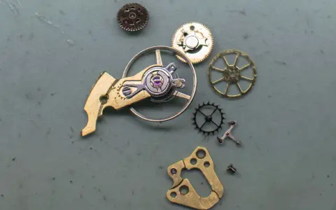 Parti di un orologio: ingranaggi, un bilanciere e altri componenti metallici disposti su una superficie grigia.