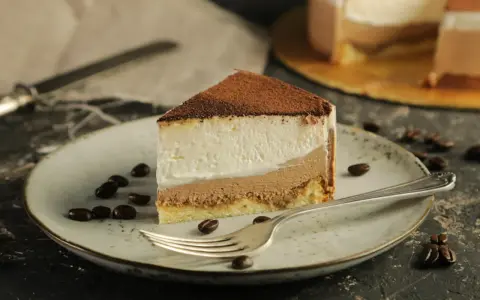 Pyszny sernik tiramisu z warstwami kremu i biszkoptu, posypany kakao. Na talerzu ziarna kawy i widelec.