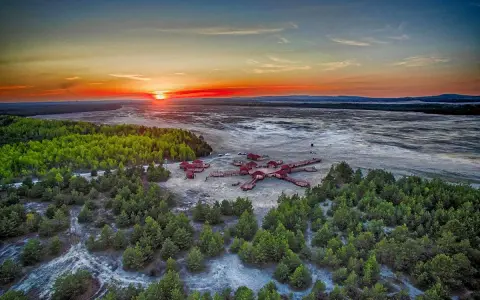 Zachód słońca nad Pustynią Błędowską. Drewniana konstrukcja z domkami przypomina rozgwiazdę na piaskowym tle, otoczona zielonym lasem.