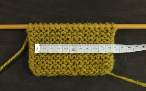Knitted swatch with a measuring tape across it, showing stitches on wooden needles.