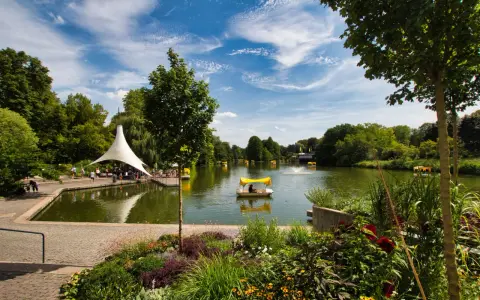 Ein sonniger Tag am Badesee Mannheim. Mehrere Tretboote mit gelben Verdecken gleiten über das Wasser, umgeben von üppigem Grün und blühenden Blumen.