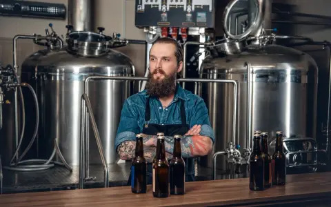 Un brasseur barbu pose devant une cuve chauffante, des bouteilles de bière sur le comptoir.