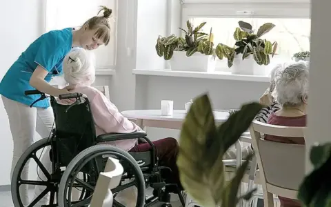 Opiekunka pomaga pacjentowi geriatrycznemu w wózku inwalidzkim. W tle siedzą inne starsze osoby.