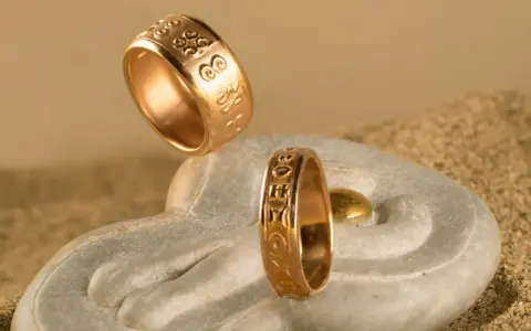 Due anelli d'oro con incisioni uniche, che mostrano diversi tipi di fedi nuziali, su una pietra a forma di mano con sabbia.
