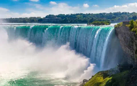 Potężny wodospad Niagara w całej okazałości. Woda spada kaskadami, tworząc mgłę. W tle drzewa i budynek.