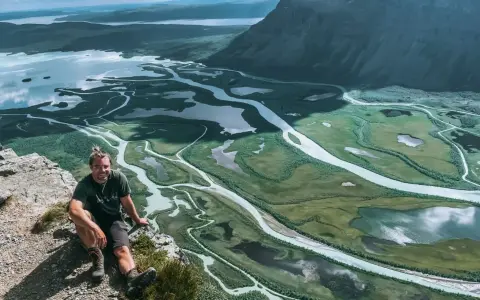 Man sitter på en klippa med en fantastisk utsikt över ett meandrande vattendrag och fjällandskap i Sarek. Aktse till skierfe!