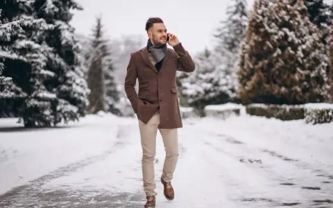 Man in stylish winter cocktail attire for men, wearing a brown coat and scarf, walks through a snowy park while on the phone.