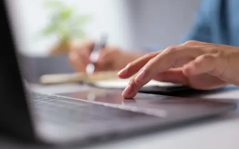 Dłoń na touchpadzie laptopa, w tle notatnik i długopis.