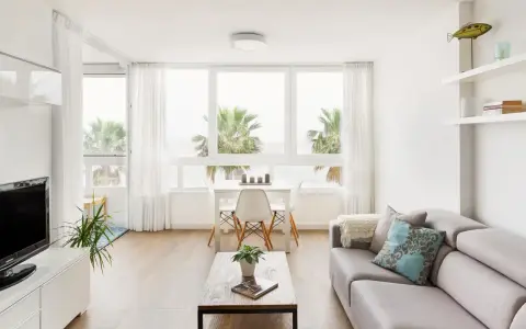 Un luminoso salón estilo nórdico con sofá gris, mesa de centro de madera y grandes ventanales con vistas a palmeras.
