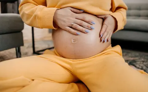 Schwangere Frau hält ihren Bauch, spürt den Druck im Unterleib. Gelber Jogginganzug, blaue Nägel.