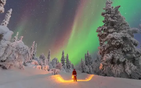 Un uomo con lanterna, vestito da Babbo Natale, ammira l'aurora boreale in Lapponia. Un paesaggio innevato e magico, perfetto per chi cerca dove andare in Lapponia.