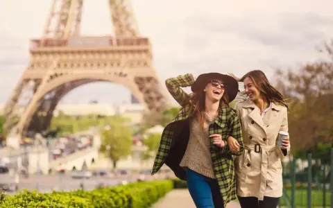 Due amiche ridono passeggiando a Parigi, con la Torre Eiffel sullo sfondo. Perfetto per capire cosa mettere in valigia per Parigi: cappotti leggeri, cappello e occhiali da sole.