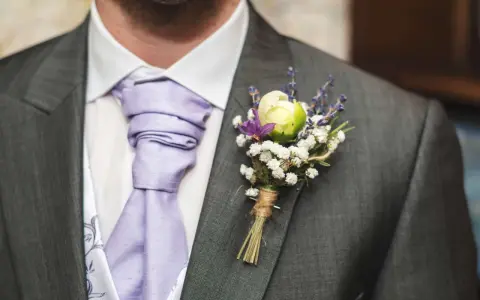 Elegancki pan młody z fioletowym krawatem i bukiecikiem kwiatów do butonierki, ozdobionym lawendą i białymi drobinkami.