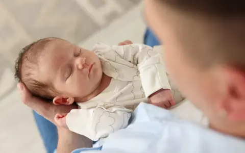 Un neonato dorme sereno tra le braccia di un genitore, cullato da suoni che ricordano i rumori bianchi, creando un'atmosfera di pace.