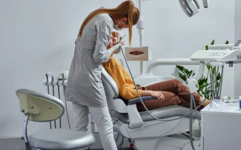 Dentystka w masce i rękawiczkach wykonuje zabieg pacjentowi siedzącemu na fotelu dentystycznym.