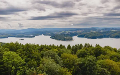Widok na jezioro otoczone lasami, z górami w tle. Na pierwszym planie bujna zieleń drzew, a dalej spokojna tafla wody i zabudowania. Nad wszystkim chmury, jakby biała góra na niebie.