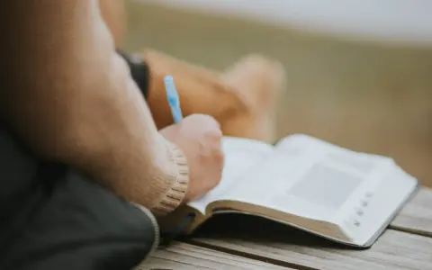 Osoba pisze w otwartej księdze, być może dziennik. W tle widać fragmenty ubrania. To moment refleksji, gdzie żal doskonały może znaleźć swoje ujście w słowach.