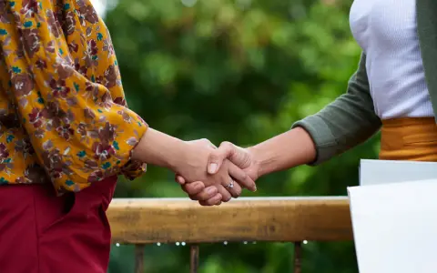 Två personer skakar hand över ett räcke utomhus. En har en blommig gul blus och röda byxor, den andra en vit topp och grön kofta.