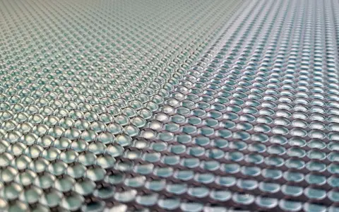 Close-up of a 3d printed lens array, with rows of tiny, clear, dome-shaped lenses reflecting light.
