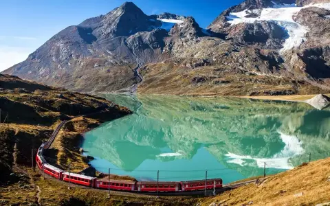 Czerwony pociąg wije się wzdłuż turkusowego jeziora u podnóża alpejskich szczytów. Idealne miejsce na trekking w alpach.