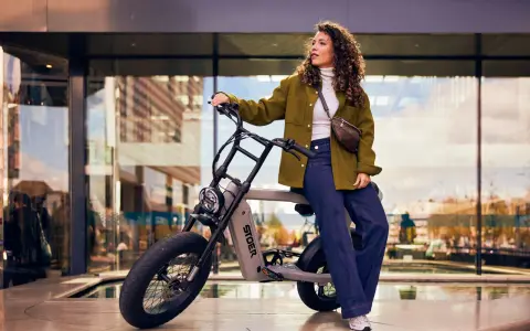 En kvinna med lockigt hår sitter på en STOER elcykel framför en modern byggnad. Hon bär en grön jacka och jeans.