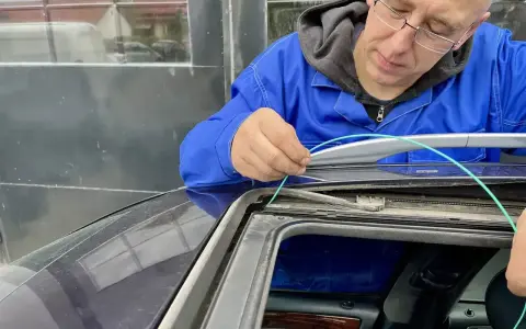 Mechanik w niebieskim kombinezonie pokazuje, jak udrożnić odpływ wody w samochodzie, używając zielonego przewodu do czyszczenia.