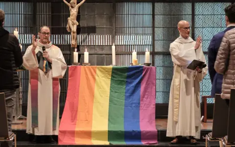 Zwei Geistliche stehen vor einem Altar mit einer Regenbogenfahne. Die Kirche feiert die gleichgeschlechtliche Ehe.