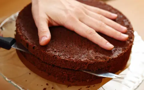 Eine Hand hält einen Schokoladenkuchen, während ein Messer ihn waagerecht durchschneidet, um den tortenboden zu teilen.