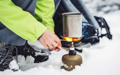 Tänd ett bensinkök i snön för att värma upp en kastrull. En person i en ljusgrön jacka och vandringskängor förbereder mat utomhus.