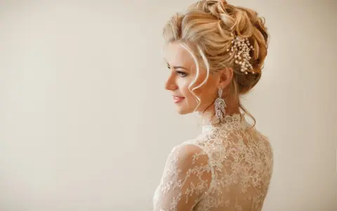 Eleganckie uczesania na wesele średnie włosy: blondynka w koronkowej sukni z perłową ozdobą i długimi kolczykami.