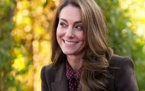 Kate Middleton sonríe con calidez, con su cabello castaño cayendo en cascada. El fondo bokeh de tonos otoñales complementa su elegante atuendo.