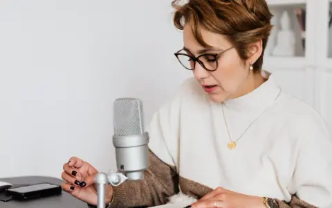 Mujer con gafas lee un libro frente a un micrófono, aprendiendo como escribir mejor.
